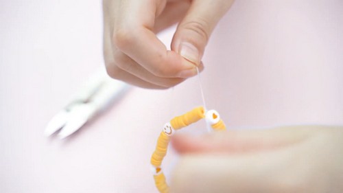 Hands pulling elastic cord through a partially strung bracelet with yellow disc beads and white smiley face beads against a pink background