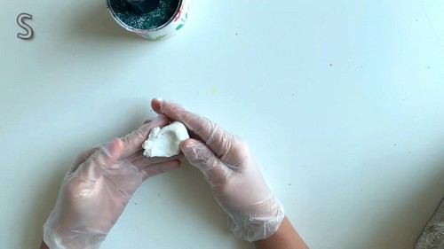 Gloved hands conditioning a piece of white polymer clay on a white work surface beside a jar of glitter.