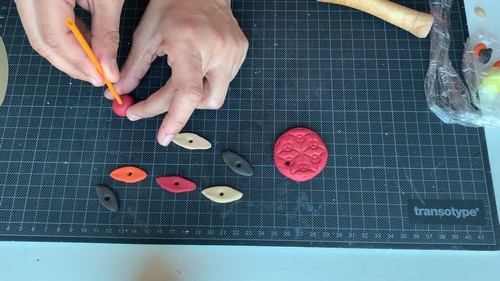 Hands using an orange piercing tool to make holes through small clay bead shapes laid out on a cutting mat.