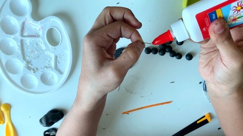 A hand applying glue with a fine tool to small black clay beads next to a paint palette and craft bottle.