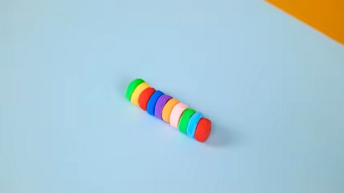 A row of flat disc-shaped clay beads in rainbow colors including green, red, blue, purple, orange, white, and red, laid in a line on a light blue surface.