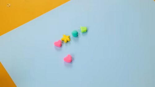 Several small unbaked clay shapes laid out on a light blue surface including two pink hearts, a yellow star, a teal circle, and a green shape with a jump ring inserted.