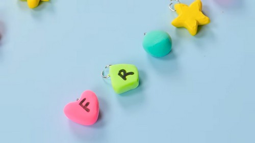 Close-up of three finished clay letter charm beads with jump rings attached, showing a pink heart stamped with F, a lime green square stamped with R, and a teal circle, with a yellow star visible above.