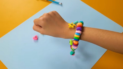 A child's wrist wearing a finished clay bead bracelet with a rainbow sequence of large disc beads and small charm shapes including a yellow star and green accent bead, with a small pink heart clay piece resting on the blue paper surface nearby.
