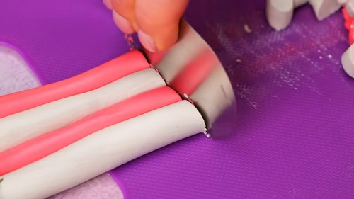 Hands slicing a layered roll of pink and white polymer clay into individual disc beads using a blade on a purple work mat.