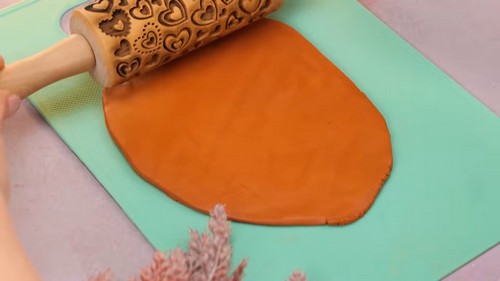 A decorative wooden rolling pin being used to flatten a large sheet of orange polymer clay on a green silicone mat.