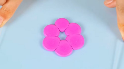 Six pink teardrop-shaped clay petal pieces arranged in a flower formation on a light blue surface, with a gap at the center where the flower middle will be placed.
