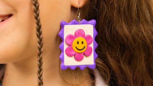 A close-up of a person wearing a large polymer clay earring shaped like a postage stamp, with a pink smiley face flower on a cream background and a purple ruffled border, hanging from a silver ear wire.