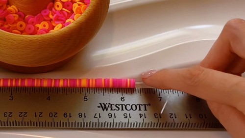 Partially strung pink and orange clay disc bead bracelet laid against a metal ruler measuring roughly seven inches beside a bowl of loose beads