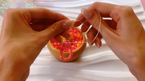 Hands tying off clear elastic cord above a wooden bowl filled with pink and orange clay heishi disc beads