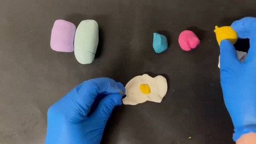 Blue-gloved hands pressing a small amount of yellow clay softener liquid onto a flattened white polymer clay piece on a dark surface, with colored clay blocks lined up in the background.