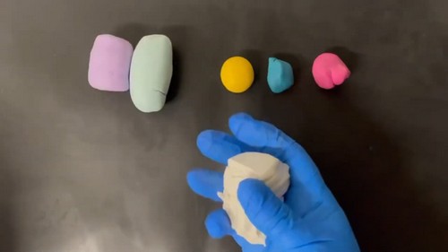 A gloved hand kneading a white clay block on a dark surface next to several unconditioned polymer clay blocks in purple, mint, yellow, blue, and pink.