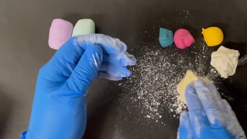 Blue-gloved hands breaking apart crumbly conditioned clay chunks over a dark surface scattered with clay dust, with softened colored blocks visible in the background.