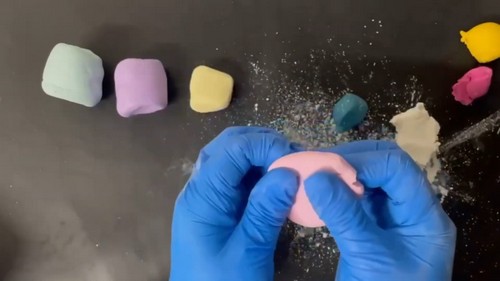 Blue-gloved hands folding and pressing a pink polymer clay block that has become pliable, with other conditioned pastel clay blocks lined up behind it.