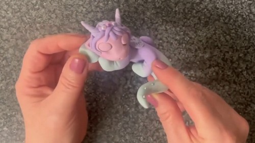 Hands holding a lavender and mint polymer clay unicorn figurine and pressing a small mint crescent tail piece with pearl beads into position.