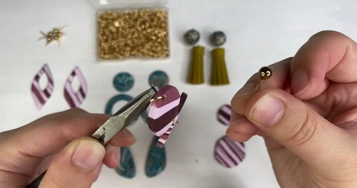 Hands using pliers to open a gold jump ring and attach it to a hole in a striped polymer clay earring piece, with gold findings and olive tassels visible in the background. 