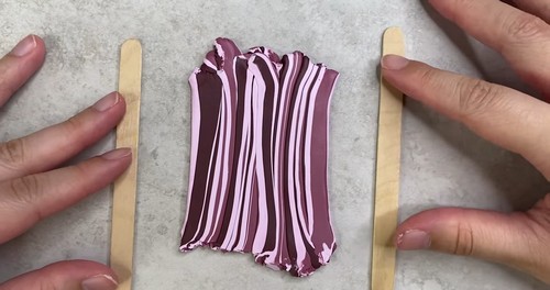 Hands pressing a folded multi-stripe pink and burgundy polymer clay cane flat between two popsicle sticks as thickness guides on a marble surface. 