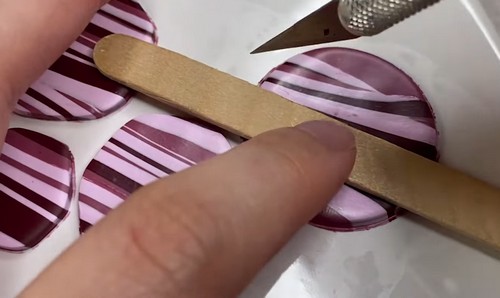 A finger and popsicle stick holding a striped pink and burgundy clay circle in place while a craft knife cuts along the edge to clean up the shape. 
