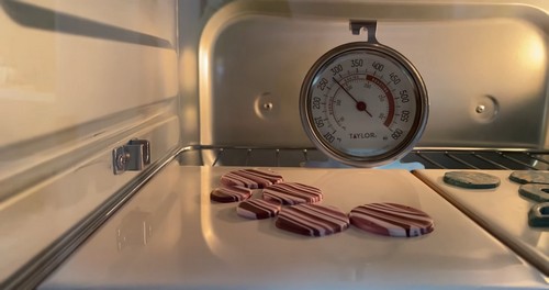 Several striped pink and burgundy polymer clay earring pieces baking on a ceramic tile inside a toaster oven with a Taylor oven thermometer visible. 