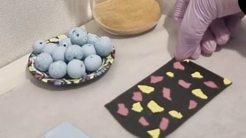 Scattered pink and yellow clay scraps arranged over a black clay base next to a dish of blue beads