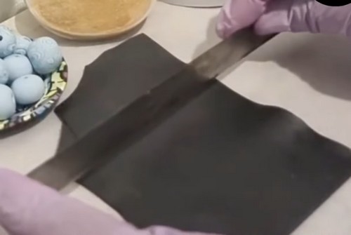 Gloved hands smoothing a rolled sheet of black polymer clay with a flat metal blade