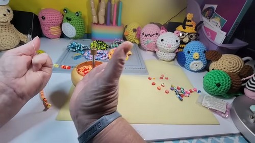 Hands holding a beaded strand with small letter and emoji beads scattered on a yellow mat beside a wooden bead spinner.