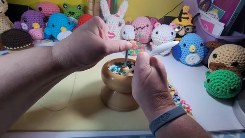 Hands picking beads from a wooden bead spinner bowl with amigurumi plush figures visible in the background.