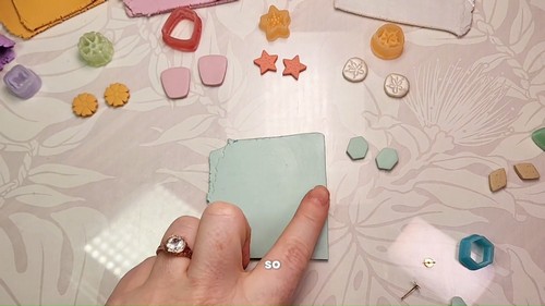 A hand pressing a sheet of mint green polymer clay flat on a patterned work surface, surrounded by small cut clay shapes in various colors and forms.