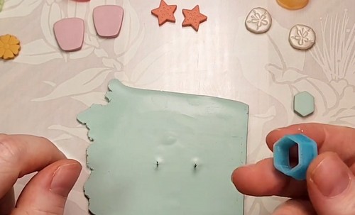 Hands pressing a small hexagon clay cutter into a mint green clay sheet, with two pin marks already visible where earring posts will be embedded before baking.