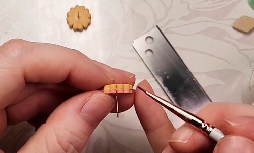 Hands pressing a gold earring post into the back of a small mustard yellow flower shaped clay piece using a needle tool to guide it into position.