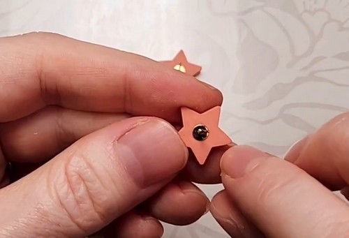 Hands holding a baked terracotta star clay earring stud and pressing a gold post pad firmly into the center of the back with a fingertip.