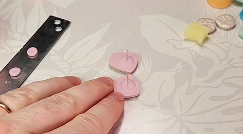 A hand steadying two unbaked pink clay stud shapes on a work surface, each with a gold earring post standing upright embedded before baking.