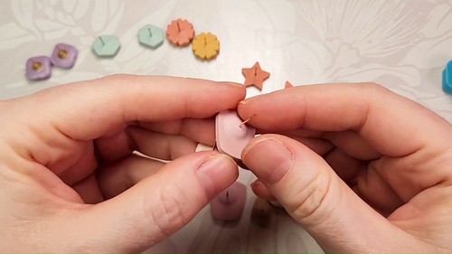 Hands bending a pink clay stud earring gently to check how securely the gold post is seated after baking, with other finished clay studs visible in the background.