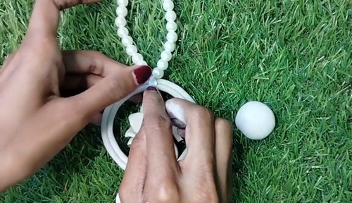 Hands attaching a pearl bead chain to a white circular clay pendant with a small clay ball beside it on grass.