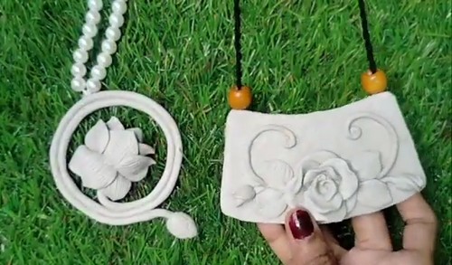 Two finished white clay necklaces side by side on grass, one with a circular lotus frame on pearls and one with a sculpted rose on a black cord.
