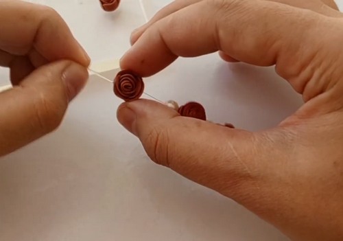 Hands threading elastic cord through a small brown clay rose bead beside a single white pearl bead during bracelet assembly.