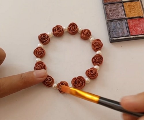A paintbrush applying color from a watercolor palette to a finished clay rose bead bracelet laid flat on a white surface.