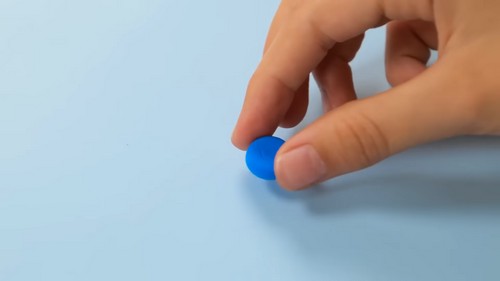 Fingers pressing a small blue clay ball into a flat disc shape against a light blue background