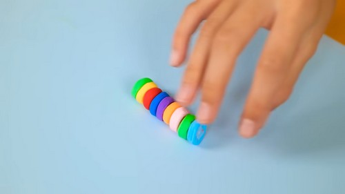 Hand pushing a row of rainbow colored clay disc beads along a blue surface showing the sequence of green, red, orange, purple, white and blue