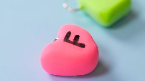 Finished pink clay heart charm with a black letter stamped on the front and a silver jump ring attached, green charm visible behind