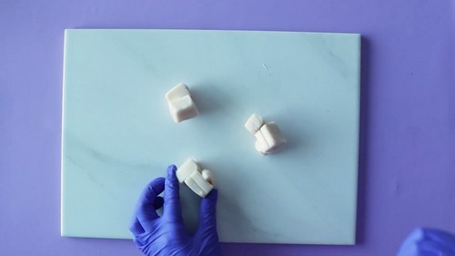 A blue-gloved hand holding a small white polymer clay block on a light blue marble tile surface with two other unconditioned white clay blocks nearby. 