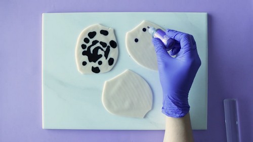 A blue-gloved hand using a dropper bottle to add dots of black alcohol ink onto two flattened white polymer clay pieces on a marble tile, with a third piece already covered in black spots. 
