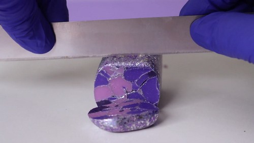 Blue-gloved hands pressing a blade down through a thick purple and silver foil-embedded polymer clay block, revealing a mosaic-like cross-section of purple tones and glitter veining. 