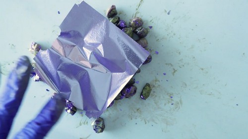A blue-gloved hand resting next to a crumpled square of purple metallic foil on a clay work surface surrounded by small clay scraps with foil fragments pressed into them. 