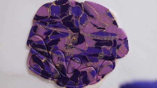 A top-down view of many thin slices of purple and gold glitter polymer clay arranged in a circular mosaic pattern on a white surface after being cut from the cane. 