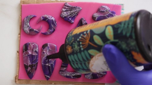 A blue-gloved hand applying liquid clay from a bottle onto several unbaked purple glitter clay earring shapes in different forms laid out on a pink foam sheet. 