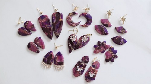 A flat lay of twelve pairs of finished purple and gold polymer clay earrings in varied shapes including fans, moons, teardrops, hearts, and crescents, all with gold hardware on a white surface.