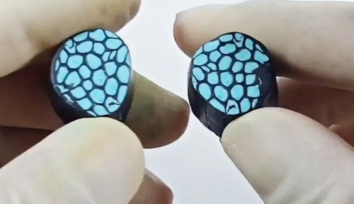 Gloved hands holding two compressed and reduced black and teal polymer clay cane cross-sections showing the irregular cellular pattern forming across the face.