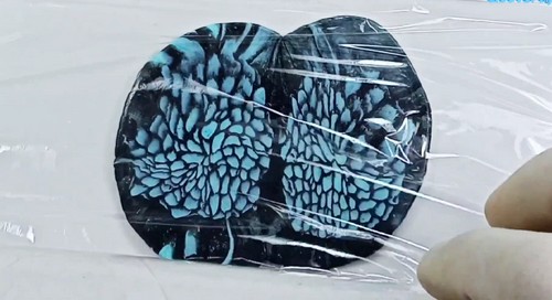 A large flattened round sheet of black and teal cane slices arranged and pressed together on a plastic-wrapped white surface, showing the full chrysanthemum-like petal pattern.