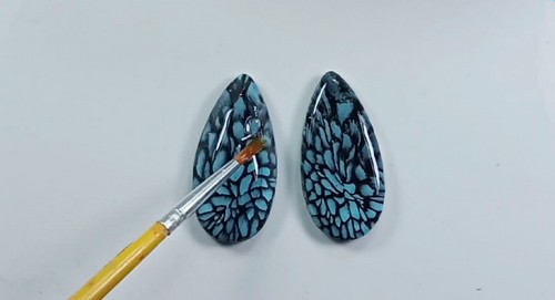 A fine paintbrush applying resin or gloss varnish onto two baked teardrop-shaped black and teal patterned polymer clay earring pieces on a white surface.